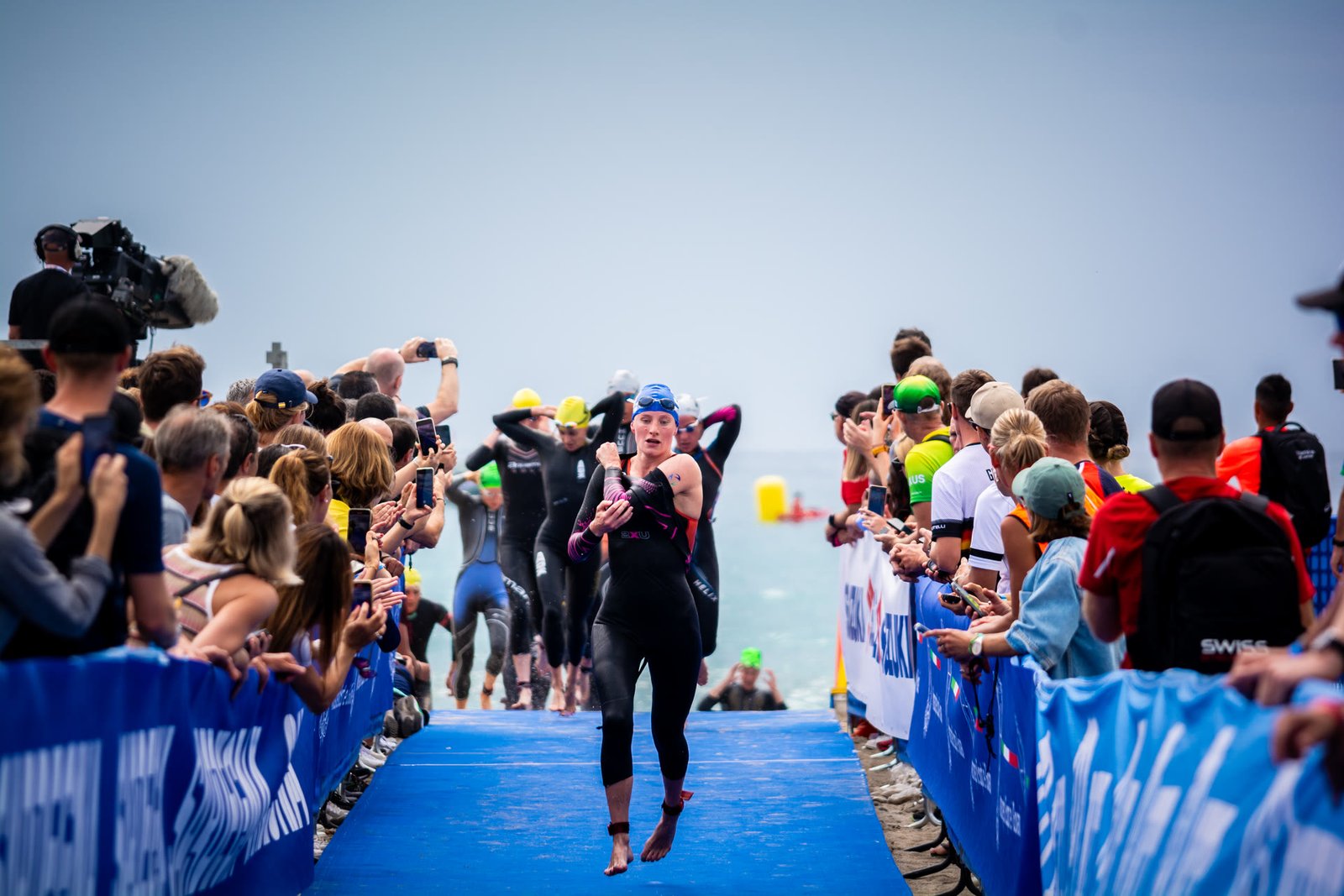 Atleta che esce dall'acqua sul tappeto di gara mentre il pubblico si sporge dalle transenne per riprenderlo