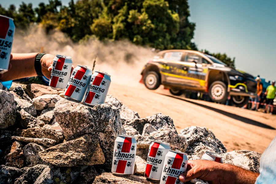 Lattine dello sponsor appoggiate sulle rocce con la vettura da rally che passa sullo sfondo