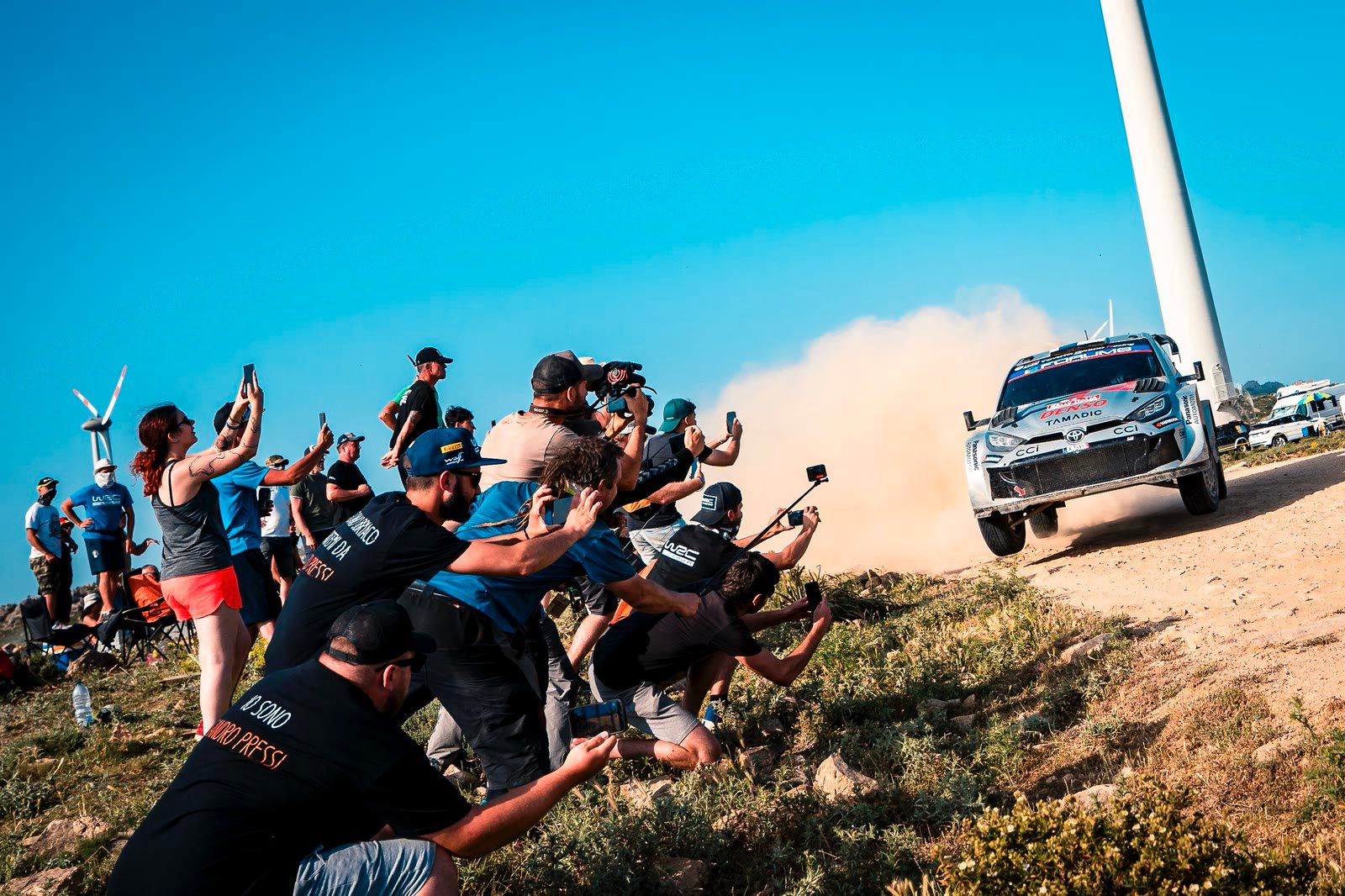 Vettura del Mondiale Rally in derapata nella polvere mentre il pubblico si sporge a fotografarla