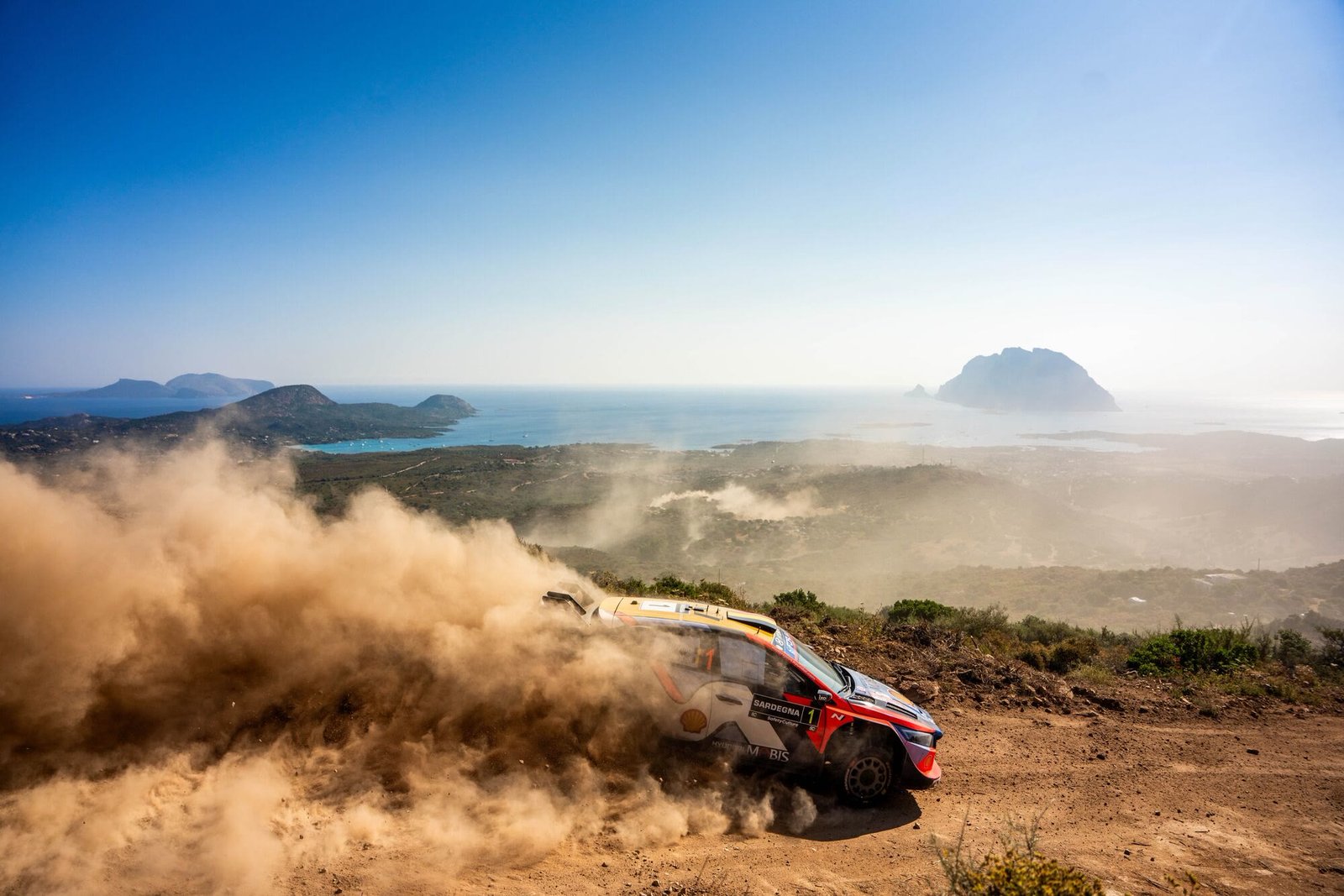 Vettura da rally che solleva la polvere sul crinale della Costa Smeralda, con il mare e le isole sullo sfondo