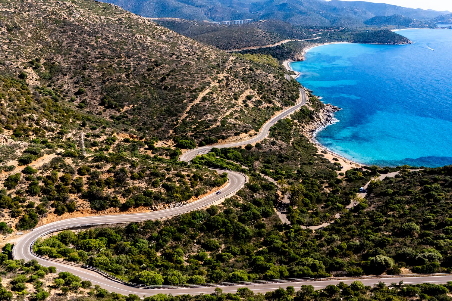 Strada costiera che scende a tornanti verso una cala di mare turchese, vista dall'alto